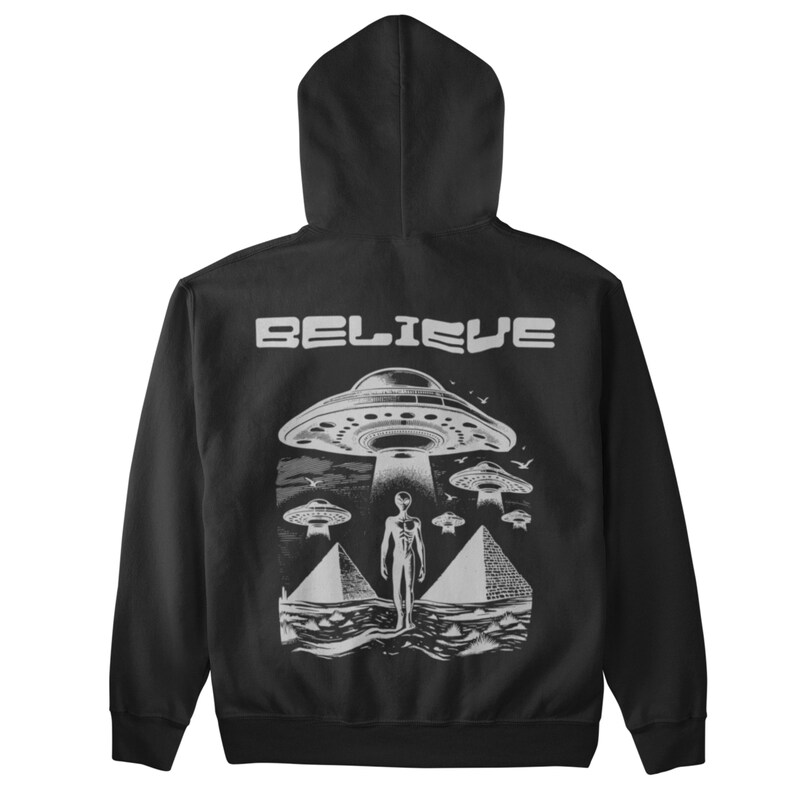 Pode incluir: Camisola de moletom com capuz preta com uma estampa gr&aacute;fica branca de um OVNI e a palavra "BELIEVE" nas costas.