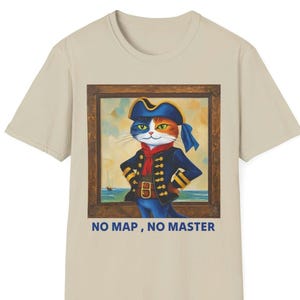以下が含まれることがあります： ベージュ色のTシャツ。海賊の格好をした三毛猫が描かれた額縁入りの絵がプリントされています。青い帽子とジャケットを着ています。絵には海の背景があり、下には「NO MAP, NO MASTER」の文字があります。