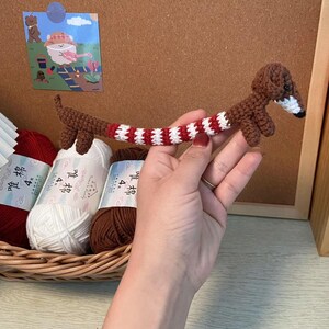 May include: A brown and white crocheted dachshund with red and white striped legs. The dachshund is being held in a hand with a red bracelet.