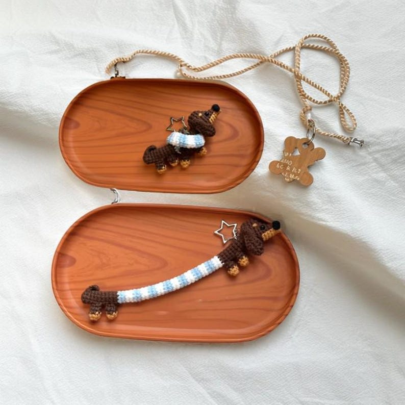 May include: Two crocheted dachshund dog keychains on wooden trays. One dachshund is wearing a blue and white scarf and the other has a blue and white striped tail. Both have a silver star charm.