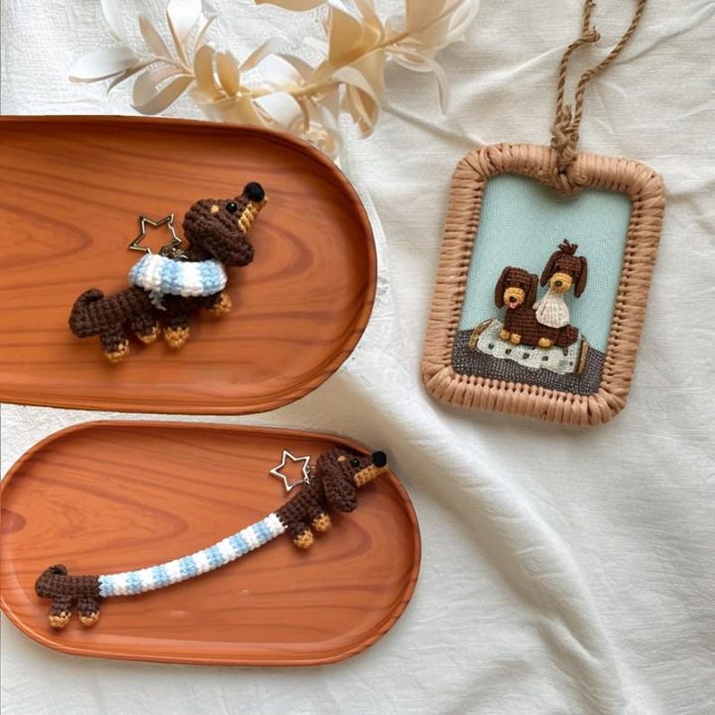 May include: Two crocheted dachshund dog keychains with blue and white striped accents. One keychain is a single dachshund, the other is a pair of dachshunds sitting together. The keychains are displayed on wooden trays.