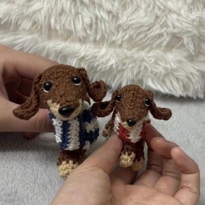 May include: Two small brown crocheted dachshund dogs wearing sweaters. The dachshund on the left is wearing a blue and white striped sweater. The dachshund on the right is wearing a red and white striped sweater.