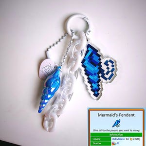 May include: A keychain with a blue iridescent seashell pendant, a pixelated blue wing charm, and a white chain. A small pink heart-shaped tag is attached. The keychain includes the text "Moz Hiar since 198".