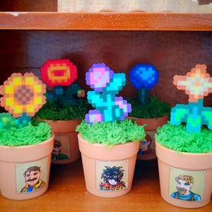 Peut inclure: Une collection de cinq petits pots en terre cuite, chacun avec un motif de fleur pixélisée différent. Les fleurs sont de différentes couleurs, dont le jaune, le rouge, le bleu et le rose. Chaque pot a une texture verte semblable à de l'herbe et une illustration de personnage.