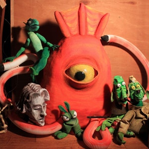 Könnte beinhalten: Eine Gruppe bunter Puppen, darunter ein rotes Monster mit einem großen Auge, ein grünes Eidechsen und zwei grüne Kreaturen mit langen Nasen. Die Puppen sind auf einer Holzoberfläche angeordnet.