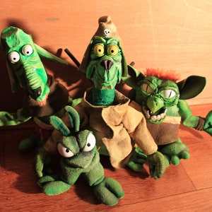 Może przedstawiać: Cztery zielone pluszowe marionetki z dużymi oczami i szerokimi uśmiechami. Marionetki są wykonane z filcu i mają różne faktury. Wszystkie noszą różne kostiumy i akcesoria.