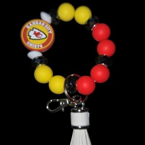 May include: A beaded wristlet keychain featuring the Kansas City Chiefs logo. The wristlet includes yellow, red, white, and black beads, a silver clasp, and a white tassel. The Chiefs logo is on a round, red and yellow charm.