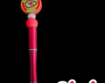 May include: Red pen with a Kansas City Chiefs logo on the top. The logo is red, yellow, and white with rhinestones around the edge. The pen is red with a silver tip.