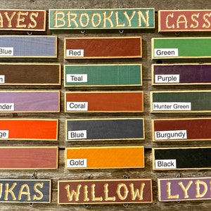 May include: A set of 12 wooden signs with various names and colors. The signs are painted in colors like light blue, red, brown, teal, purple, lavender, coral, hunter green, orange, blue, burgundy, pink, gold, and black. The names on the signs are Hayes, Brooklyn, Cassie, Lukas, Willow, and Lydia.