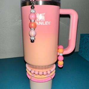 Könnte beinhalten: Ein rosa-pfirsichfarbener Stanley-Becher mit Griff, verziert mit einem Perlenanhänger und Armbändern. Der Becher hat das Stanley-Logo und ein Farbverlaufsschema. Zusätzliche Perlenarmbänder sind an der Basis gestapelt.