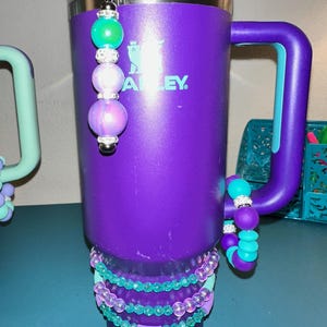 Może przedstawiać: Fioletowy kubek Stanley z uchwytem i koralikowym urokiem. Urok ma zielone, fioletowe i srebrne koraliki. Koralikowe bransoletki w odcieniach fioletu i turkusu zdobią kubek.