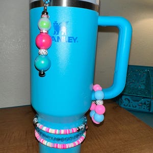 May include: A turquoise blue stainless steel travel mug with a handle. The mug has a clear lid and a straw. The mug is decorated with a beaded charm and a stack of colorful bracelets.