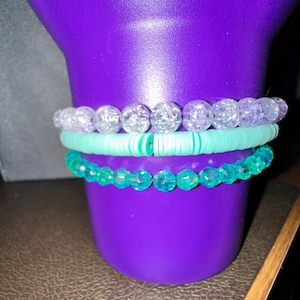 Może przedstawiać: Trzy bransoletki ułożone na fioletowym tle. Górna bransoletka wykonana jest z przezroczystych, błyszczących koralików. Środkowa bransoletka wykonana jest z jasnoniebieskich koralików. Dolna bransoletka wykonana jest z ciemnoniebieskich koralików.