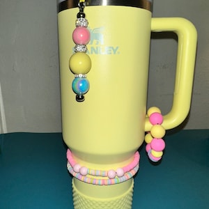 May include: A yellow Stanley tumbler with a handle, adorned with a beaded charm and bracelets. The charm features pink, yellow, and blue beads. Additional bracelets and beads in pink, yellow, and pastel colors are attached to the tumbler.