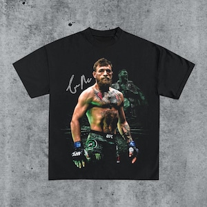 May include: Black t-shirt featuring a color image of a man with tattoos, a beard, and a signature in white. The man is wearing gloves and shorts with the UFC logo. A faded image of a man is in the background.