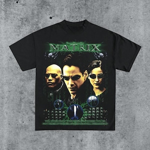 May include: Black t-shirt featuring a graphic from the movie "The Matrix." The design includes the film's title in green, along with images of the main characters. The shirt has a crew neck and short sleeves.