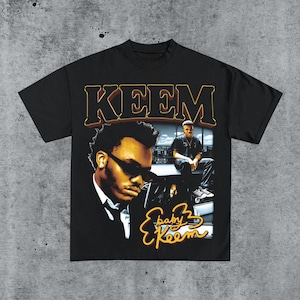 May include: Black t-shirt featuring a vintage-style graphic of a man wearing sunglasses, with the name "KEEM" in large gold lettering. The graphic also includes the words "baby Keem" in a stylized font, and a photo of the man sitting.