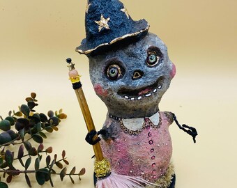 May include: A whimsical witch figurine made of paper mache, wearing a black pointed hat with gold stars and a pink dress with gold accents. She holds a broom with pink feathers and a gold handle.