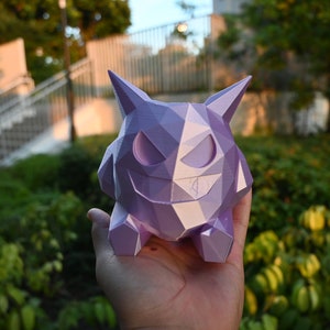 Puede incluir: Una figura de Pokemon Gengar de tipo Fantasma, impresa en 3D de baja poli, de color morado. La figura se sostiene en la mano de una persona.
