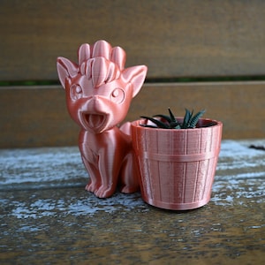 Puede incluir: Una maceta impresa en 3D con forma de animal de dibujos animados, con una pequeña maceta de estilo cubo de madera al lado. El animal es de color marrón rojizo, con una cara sonriente y orejas grandes. La maceta contiene pequeñas plantas verdes.