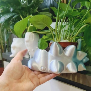 May include: A white ceramic planter shaped like a spiky creature with a light blue glaze. The planter is holding a small green plant.