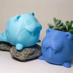 May include: Two ceramic animal planters, one light blue and one blue, shaped like a cartoon animal with a wide mouth and large eyes. The blue planter has a small succulent plant growing inside.