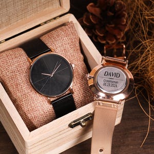 Könnte beinhalten: Zwei Uhren in einer Holzkiste. Eine Uhr hat ein schwarzes Zifferblatt und ein schwarzes Metallband. Die andere Uhr hat ein goldenes Zifferblatt mit dem Text "DAVID  Groomsman 06.19.2024" und ein goldenes Metallband.