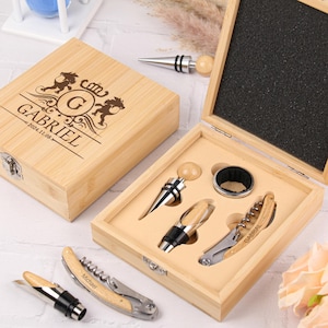 Puede incluir: Una caja de madera con una inscripción personalizada "Gabriel 2021.11.08" y un escudo con la letra "G" en su interior. La caja contiene un sacacorchos, un tapón de botella y un corta cápsulas. La caja está hecha de bambú y tiene un acabado de madera natural.