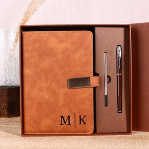 May include: A brown leather journal with a silver clasp and the initials "M K" printed on the front. The journal is packaged in a brown gift box with a matching pen and a silver pen.