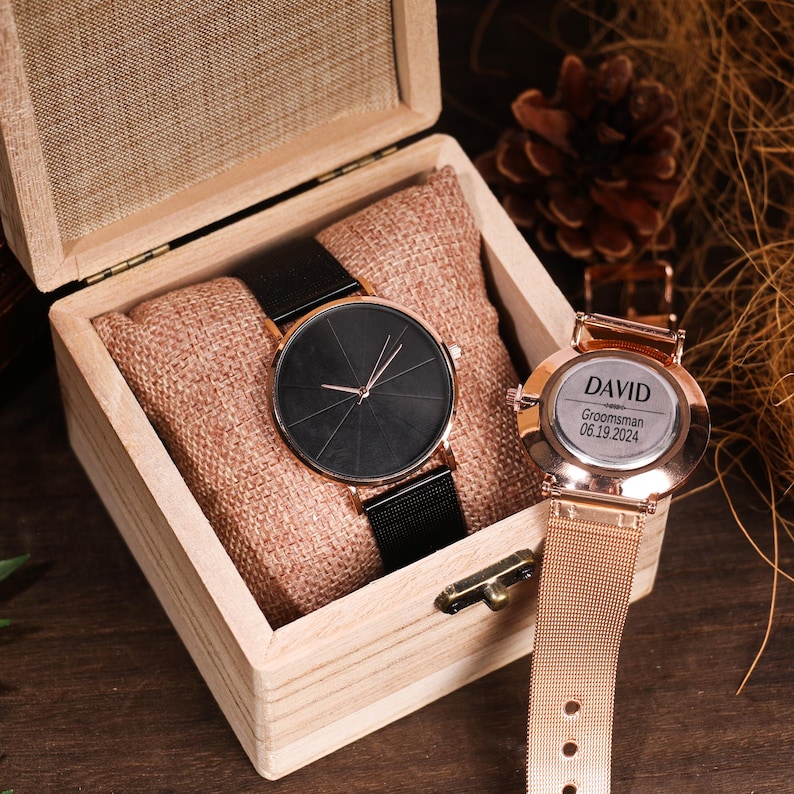 May include: Two watches in a wooden box. One watch has a black face and a black metal band. The other watch has a gold face with the text "DAVID  Groomsman 06.19.2024" and a gold metal band.
