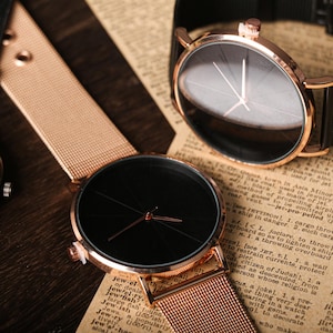 May include: Two wristwatches with black faces and gold-tone bands. The watch on the left has a mesh band and the watch on the right has a black leather band.