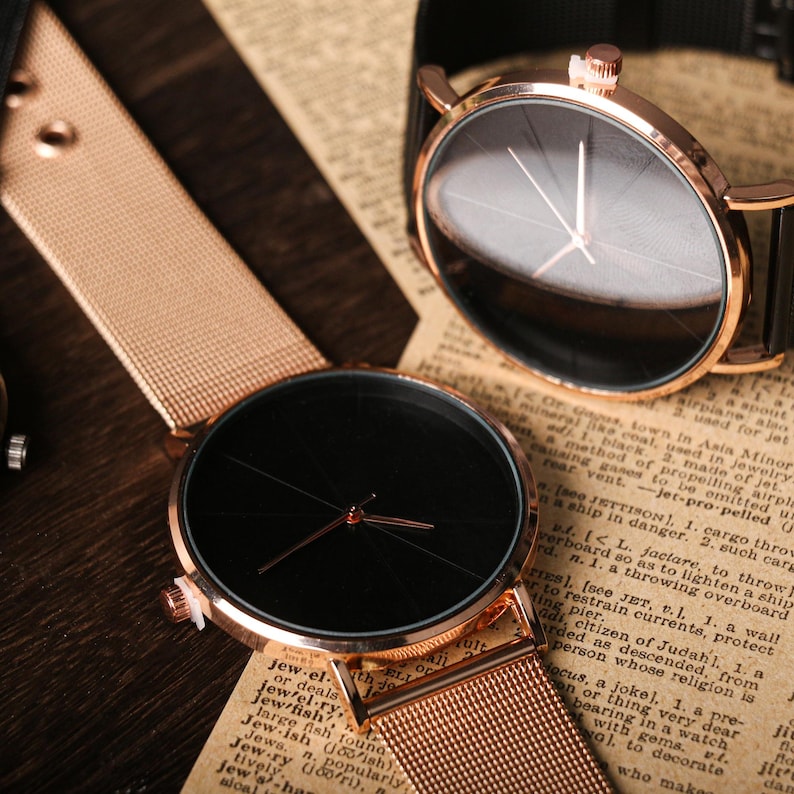 May include: Two wristwatches with black faces and gold-tone bands. The watch on the left has a mesh band and the watch on the right has a black leather band.