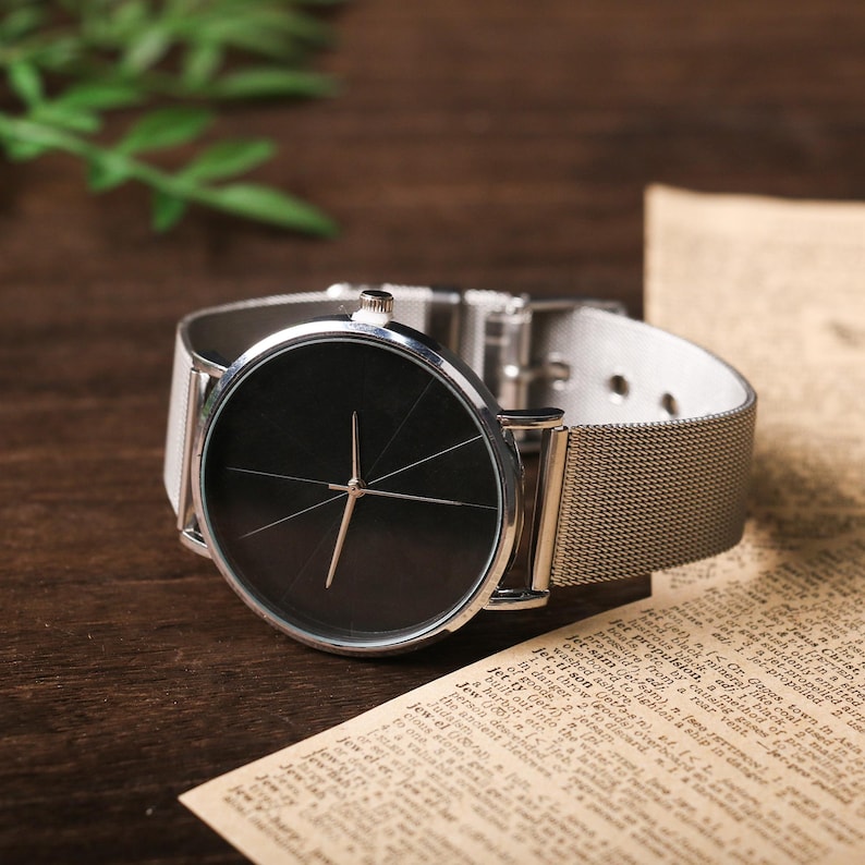 May include: A silver watch with a black face and a silver mesh band. The watch has a simple design with a single hand and no numbers.