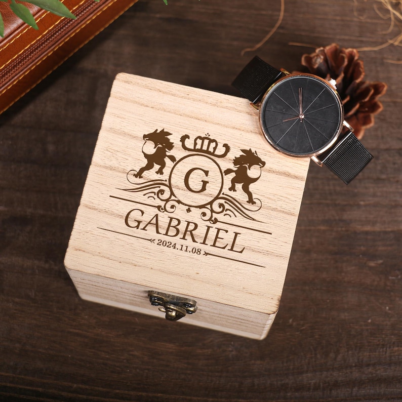 May include: A wooden box with a personalized inscription that reads "Gabriel 2024.11.08" and features a crest with the letter "G" surrounded by two lions. A black watch with a black face and a metal band is placed on top of the box.