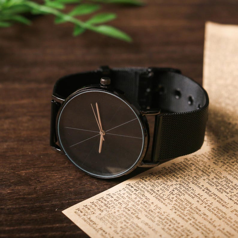 May include: A black wristwatch with a black mesh band and a round black face. The watch has a simple design with a thin silver hour and minute hand.