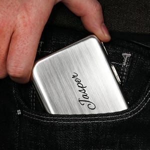 May include: A rectangular, brushed silver metal cigarette case featuring the name Jasper engraved in a black cursive script on the front surface. The case has rounded corners and a metallic latch mechanism visible on the side edge.