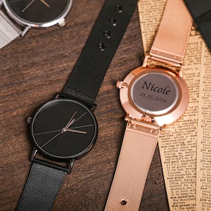 May include: Two wristwatches with metal mesh bands. One watch has a black face with silver hands and the other has a rose gold face with the text "Nicole 05.25.2024" engraved on it.