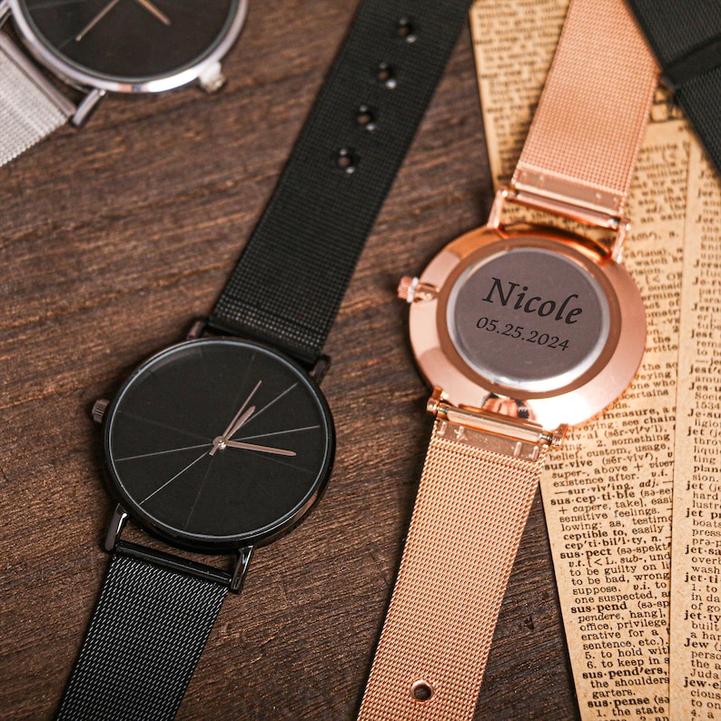 May include: Two wristwatches with metal mesh bands. One watch has a black face with silver hands and the other has a rose gold face with the text "Nicole 05.25.2024" engraved on it.