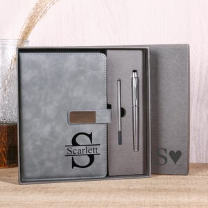 Puede incluir: Un juego de cuaderno y bolígrafo gris en una caja de regalo. El cuaderno tiene un diseño personalizado con la letra "S" y el nombre "Scarlett". El juego incluye un bolígrafo y un lápiz óptico. La caja también es gris.
