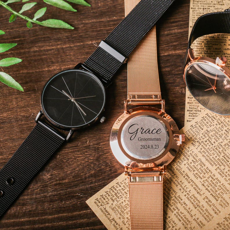 May include: Two wristwatches with mesh bands. One watch has a black face and a black band. The other watch has a gold face and a gold band. The gold watch has the text "Grace Groomsman 2024.8.23" engraved on the back.