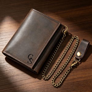 May include: A dark brown leather bifold wallet featuring a smooth matte finish and the embossed letters GS. The wallet is attached to a long, antique brass-toned metal chain that connects to a matching leather belt loop strap with a brass snap button.