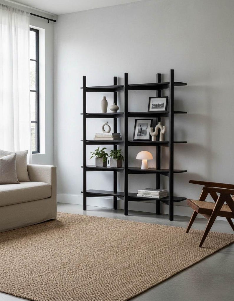 May include: A black bookshelf with multiple shelves displays decorative items like vases, framed pictures, and a small lamp. The room features a beige rug, a beige sofa, and a wooden chair, creating a minimalist interior design.