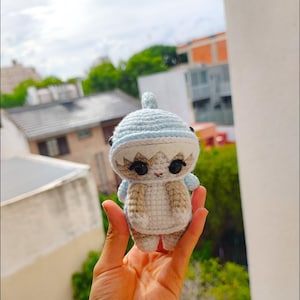 Puede incluir: Un juguete de peluche tejido a crochet con forma de tiburón que lleva un gorro azul y blanco. El juguete tiene ojos negros y una boca blanca.