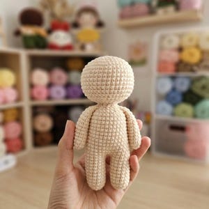 Puede incluir: Una base de muñeco de ganchillo beige que presenta una cabeza redonda, un torso, dos brazos y dos piernas. El muñeco está elaborado con un patrón de punto bajo consistente, creando una superficie texturizada en toda la figura. No tiene rasgos.