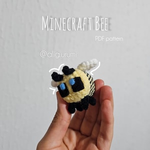 Könnte beinhalten: Eine gehäkelte Minecraft-Biene, gehalten in einer Hand, mit gelbem Körper, schwarz-weiß gestreiftem Hinterleib und schwarzem Kopf mit blauen Augen. Der Text "MINECRAFT BEE" und "PDF pattern" sind sichtbar.