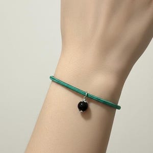May include: A green cord bracelet with a small, round, black bead charm. The charm is suspended from a silver-colored metal loop. The bracelet is worn on a wrist.