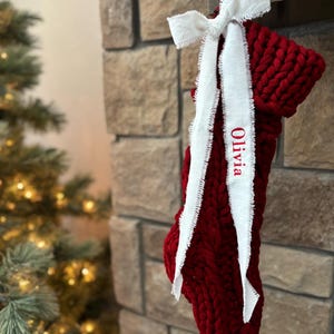 Puede incluir: Un calcetín navideño de punto grueso en color rojo intenso, colgado de una chimenea de ladrillo. Una cinta blanca con el nombre "Olivia" bordado en rojo está atada en la parte superior. Un árbol de Navidad es visible en el fondo.