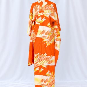 May include: A vibrant orange kimono featuring a repeating pattern of white and yellow stylized arrow feathers. The garment has long, wide sleeves, a structured collar, and a full-length silhouette displayed on a mannequin against a plain backdrop.