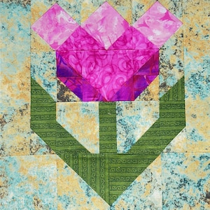 Può includere: Un blocco di trapuntatura quadrato con un motivo a tulipano stilizzato. I petali del fiore sono rosa e viola, con uno stelo e foglie verdi. Lo sfondo è un motivo screziato di blu, verde e oro.