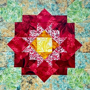 Può includere: Un colorato blocco di trapuntatura con un motivo floreale centrale. La trapunta presenta una vibrante forma di fiore rosso con un centro giallo, circondata da tessuto fantasia in rosso, bianco e motivi floreali. Lo sfondo ha quadrati verdi, blu e marroni.